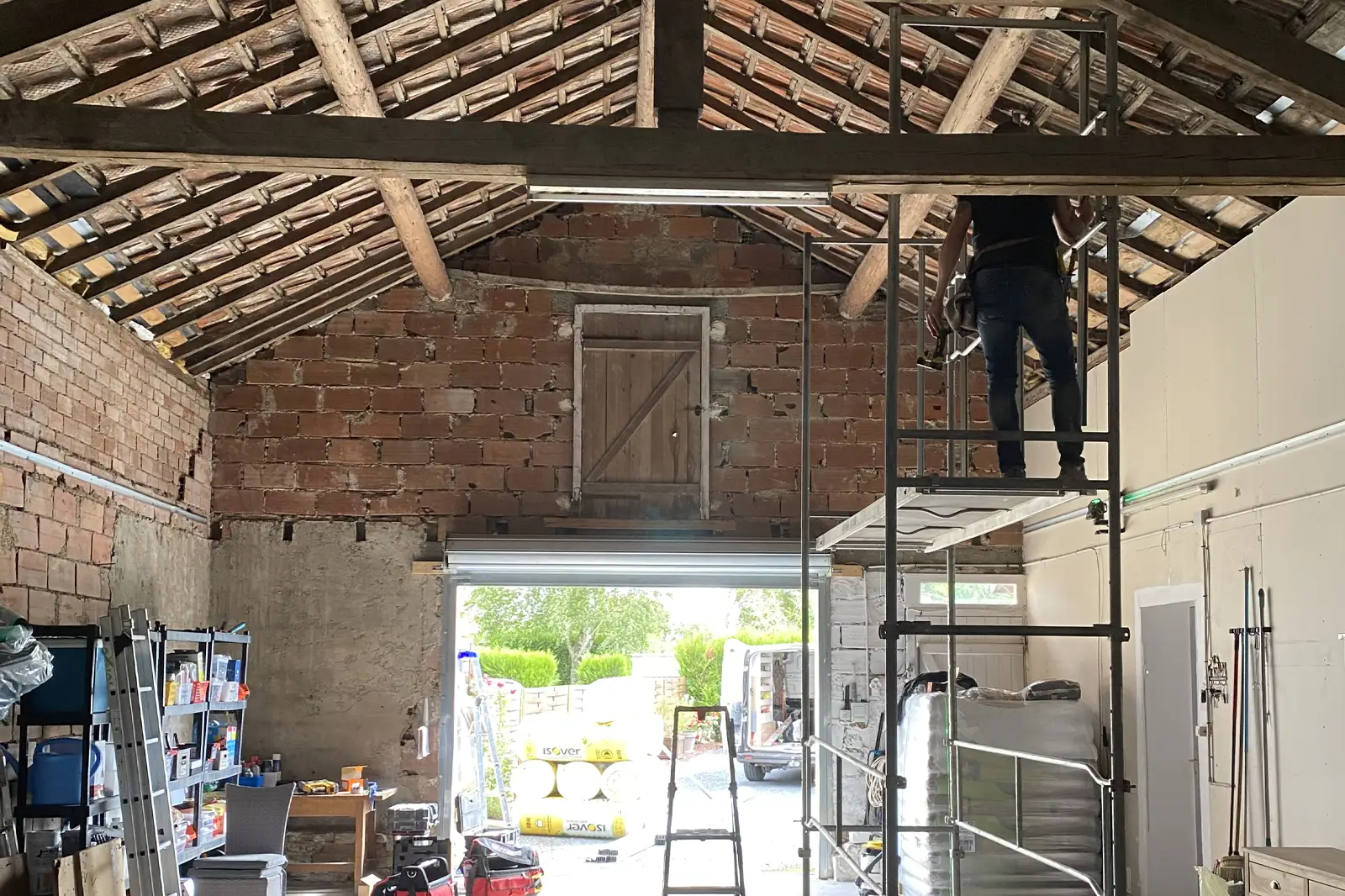 Un homme travaille sur une échelle dans un garage en rénovation avec le toit ouvert et des outils autour