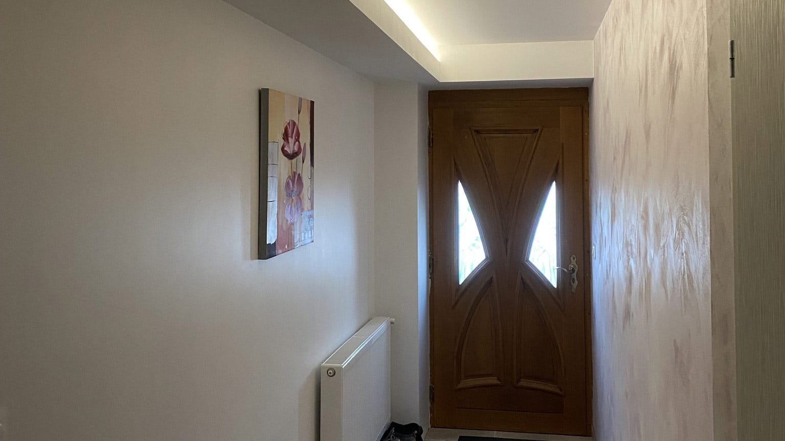 Une porte en bois avec des fenêtres en forme de losange, un tableau floral sur le mur et un radiateur blanc dans un couloir éclairé